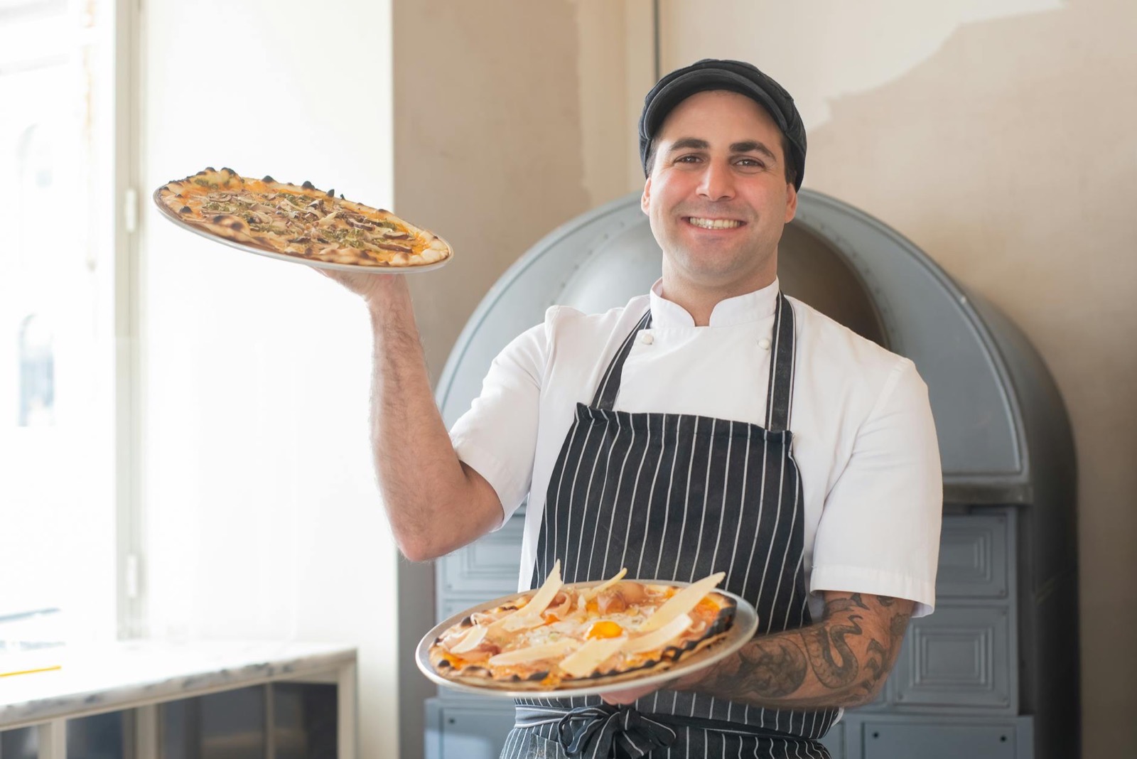 Uśmiechnięty pizzaiolo w pasiastym fartuchu trzyma dwie pizze